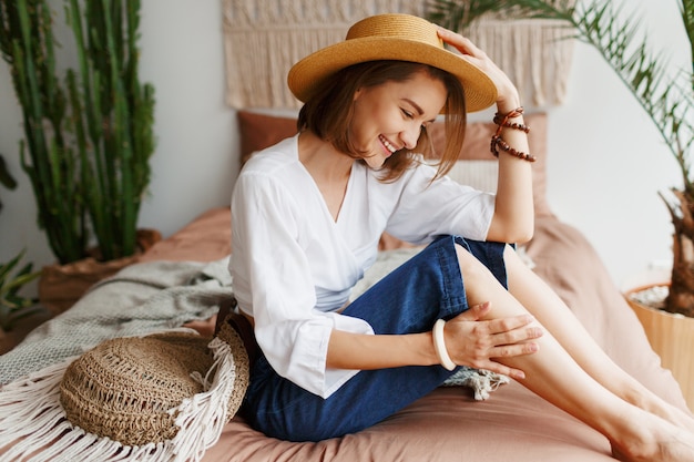 Romantic woman with candid smile sitting on bed, enjoying sunny morning in her stylish flat in boho style