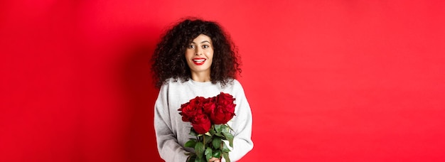 Romantic woman with curly hairstyle receive bouquet of red roses on valentines day looking tender at