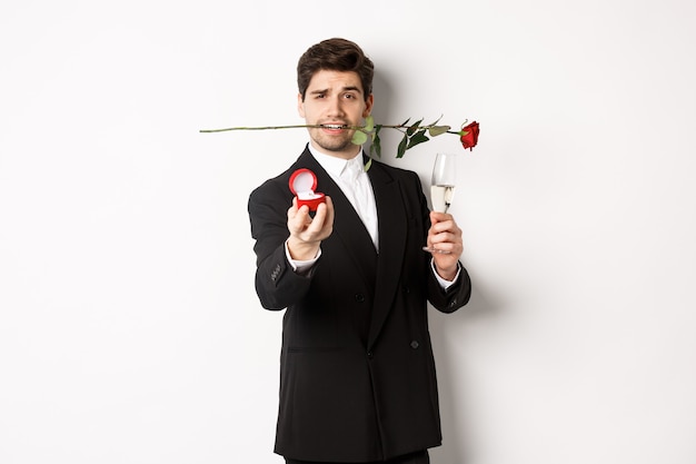 Romantic young man in suit making a proposal, holding rose in teeth and glass of champagne, showing engagement ring, asking to marry him, standing against white background