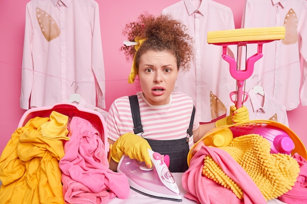 Free photo routine work and household concept. worried impressed housewife with curly hair holds mop poses near ironing board with electric iron tidies up clothes brings pile of laundry busy doing housework