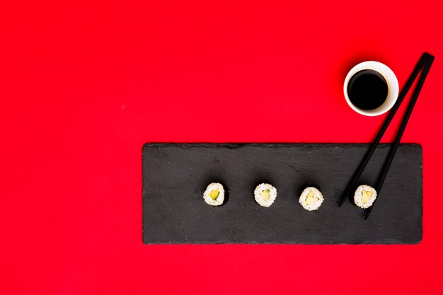 Row of asian rolls on slate plate with chopsticks and soya sauce over red backdrop