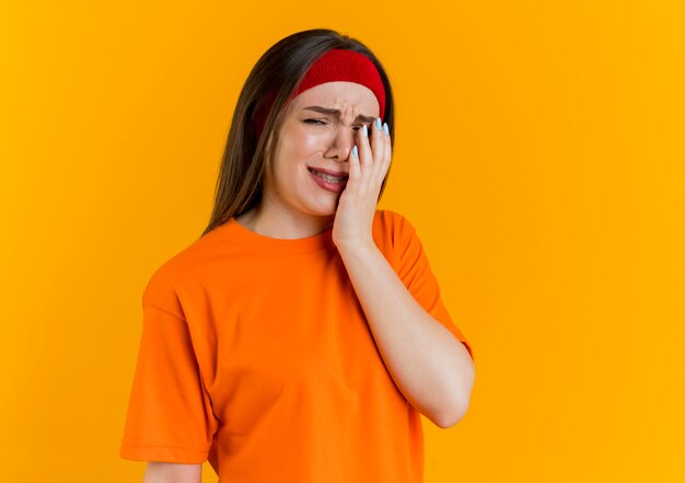 Sad aching young sporty woman wearing headband and wristbands looking putting hand on face 