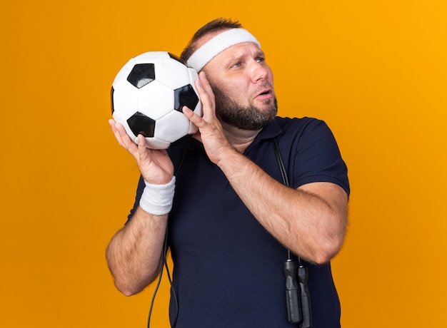 Free photo sad adult slavic sporty man with jumping rope around neck wearing headband and wristbands holding ball and looking at side isolated on orange wall with copy space