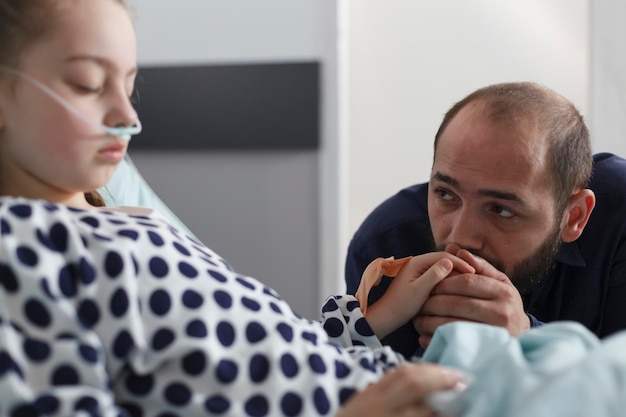 Sad anxious caring father holding sleeping ill little children hand in pediatric hospital patient ward. Sick girl using nasal tube in hospital bed while attentive parent looking at her.