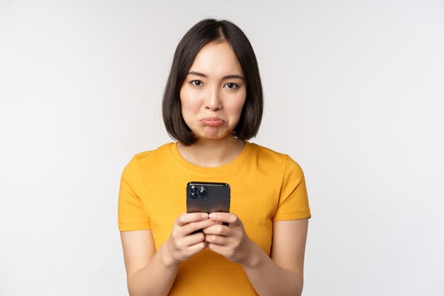 Sad asian woman holding smartphone looking upset with regret standing in yellow tshirt against white background