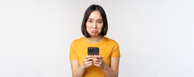 Sad asian woman holding smartphone looking upset with regret standing in yellow tshirt against white background