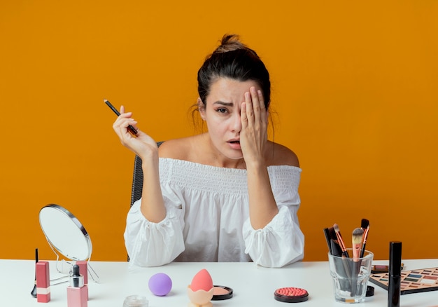 Sad beautiful girl sits at table with makeup tools closes eye with hand holding eyeliner looking isolated on orange wall