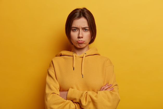 Free photo sad betrayed unhappy woman stands with arms folded, wants to cry, feels offended and lonely, wears sweatshirt, models against yellow wall, purses lower lip, thinks over serious problem