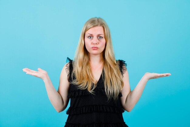 Sad bonde girl is looking at camera by opening wide her arms on blue background