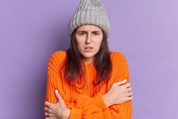 Sad brunette millennial girl with dark hair dressed in knitted wear embraces herself feels cold trembles and looks dissatisfied.