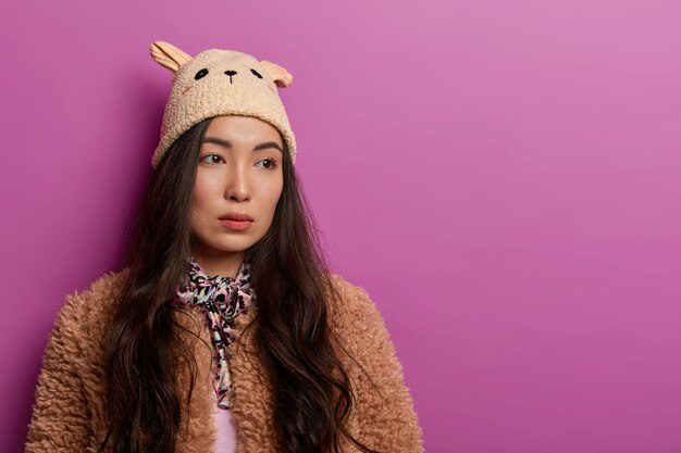 Sad brunette woman turns from camera, has seious thoughtful expression, wears hat and coat, feels lonely and discontent