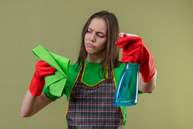 Sad cleaning young girl wearing uniform in red gloves holding cleaning spray looking at rag on her hand on isolated green background