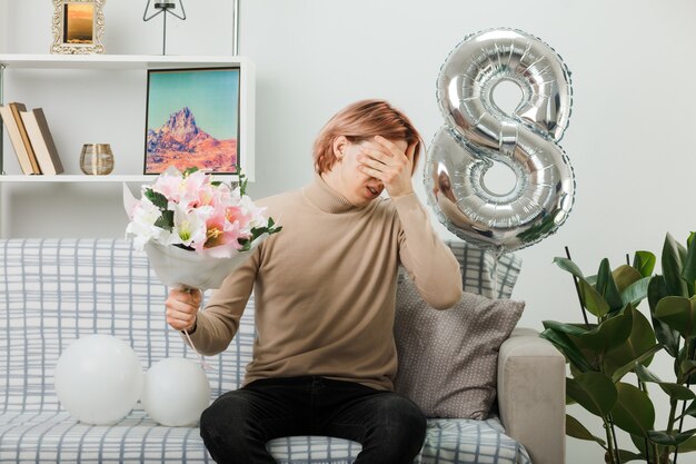 Sad covered face with hand handsome guy on happy women day holding bouquet sitting on sofa in living room