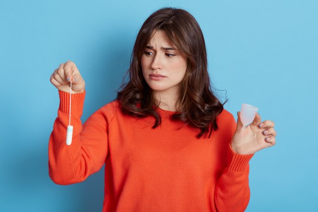 Sad dark haired female wearing orange sweater holding in hands women's hygienic products