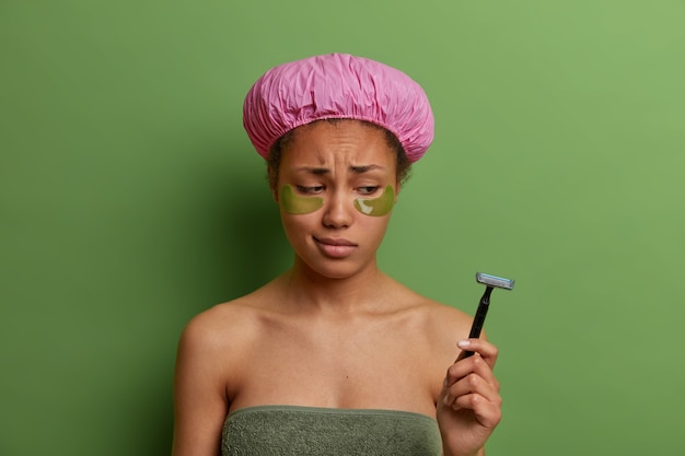 Free photo sad dejected woman looks at razor, applies collagen patches, has eye skin rejuvenation treatment, wears bathing cap after taking shower, doesnt want to shave legs, isolated on green wall