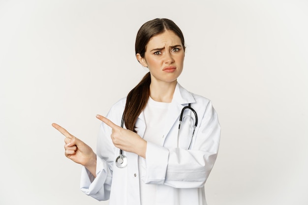 Sad and disappointed female doctor frowning frustrated pointing fingers and complaining standing in ...