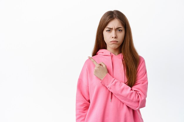 Sad and disappointed girl points left aside, frowns and looks jealous, complains on unfair thing, standing displeased against white wall