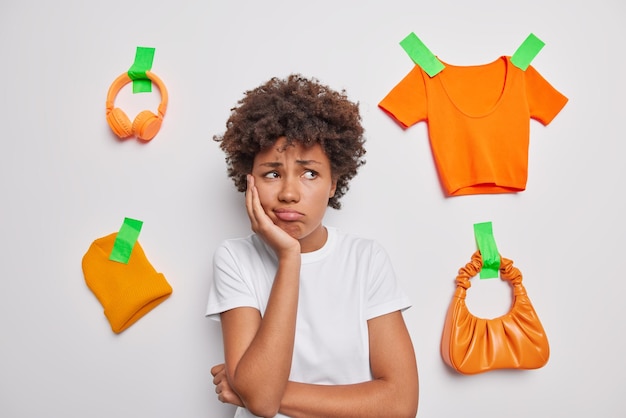 Free photo sad displeased curly haired young woman looks away unhappily dressed in casual t shirt isolated over white background with plastered orange headphones hat t shirt and bag being deep in thoughts