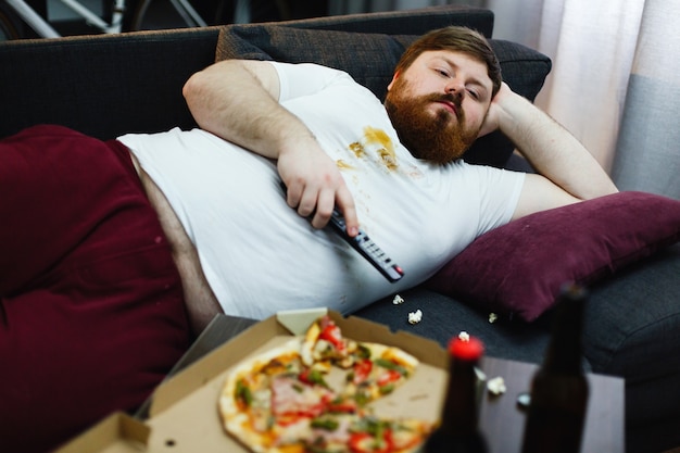 Sad fat man lies on the sofa and watches TV