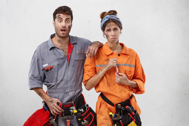 Free Photo sad female carpenter holds screwdriver, wears tool belt on waist, stand next to handyman with shocked expression