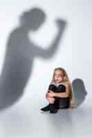 Free photo sad and frightened little girl with bloodshot, bruised eyes crying scared of shadow on the wall.