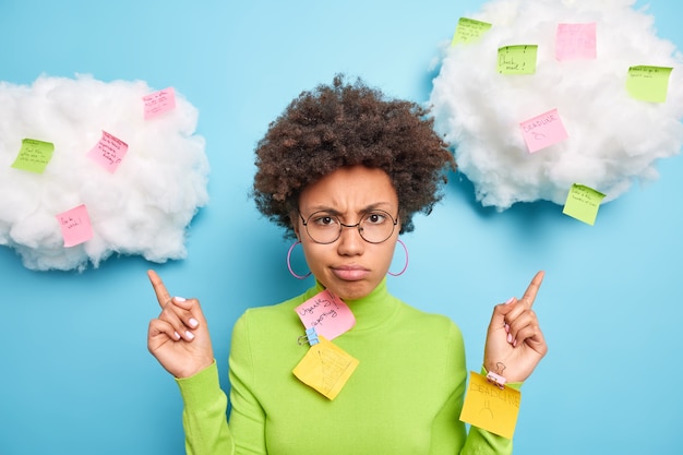 Free photo sad frustrated afro american woman point above on clouds with sticky notes written tasks to do busy as has much work wears round spectacles and green turtleneck isolated over blue wall