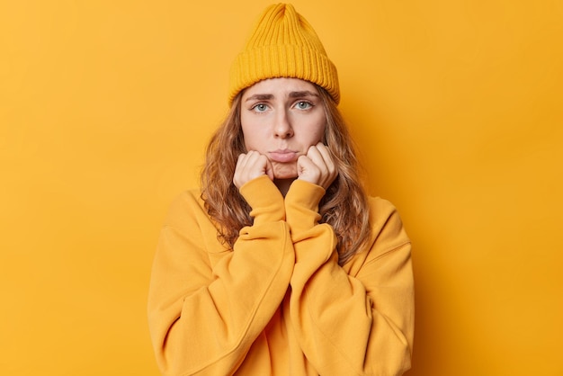 Sad frustrated woman keeps hands under chin purses lips looks discontent has displeased facial expression wears hat and jumper isolated over yellow background. Negative emotions and feelings