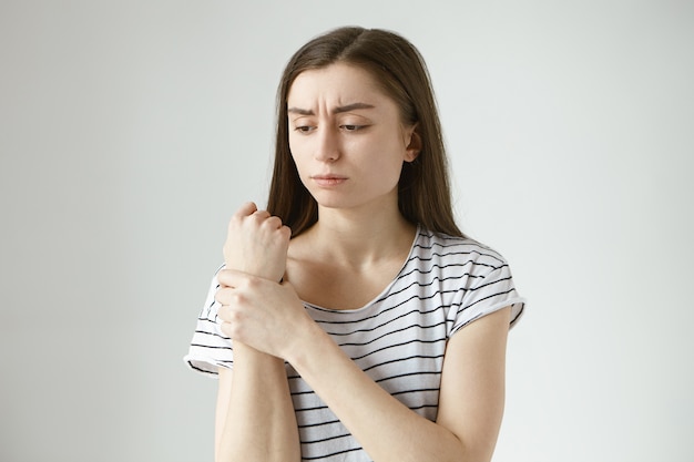  sad frustrated young woman in striped top frowning, holding hand on her aching wrist, massaging pain area, having painful facial expression, suffering from joint pain, arthritis or gout