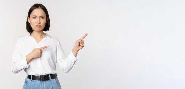 Sad and gloomy asian woman looking disappointed complaining at banner or advertisement pointing fingers right at promo and frowning upset white background