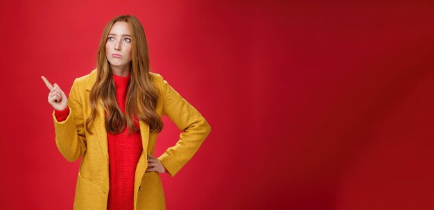 Free photo sad and gloomy cute ginger girl in s wearing trendy yellow coat holding hand on waist pouting lookin