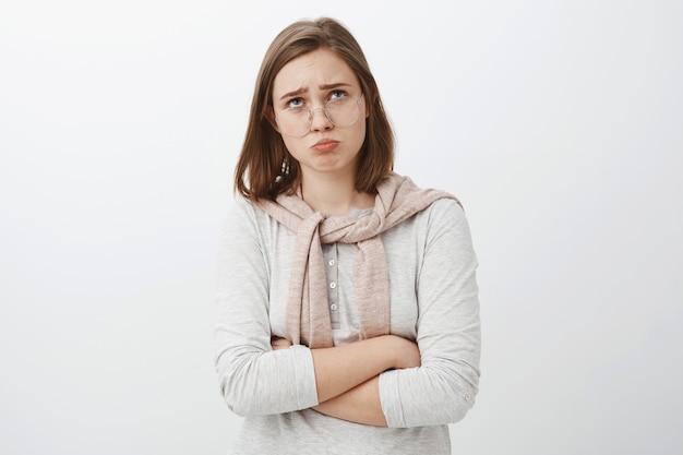 Free photo sad gloomy offended silly girl in glasses and cute outfit folding lips, pouting frowning and looking up while complaining being offended whining over cruel and unfair life over gray wall