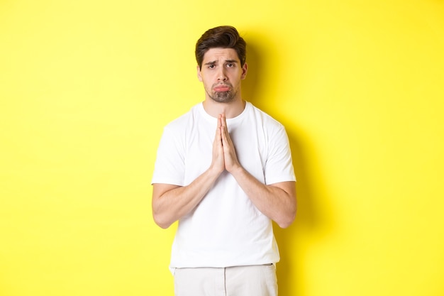 Free photo sad guy begging for something, sulking and asking for favour, saying sorry, standing over yellow background.