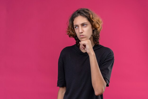 Sad guy with long hair in black t-shirt put hand under his chin on pink wall