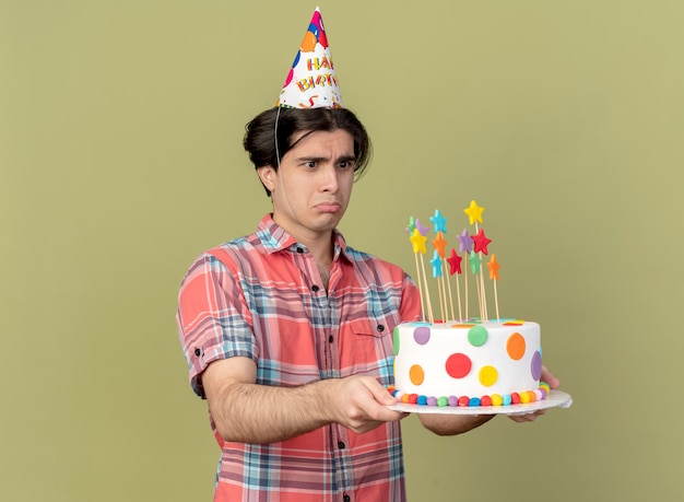 Sad handsome caucasian man wearing birthday cap holds and looks at birthday cake 