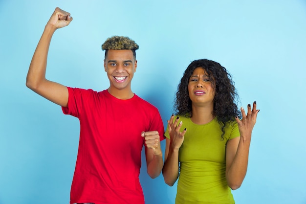 Sad and happy feelings. Young emotional african-american man and woman in casual clothes posing on blue background. Beautiful couple. Concept of human emotions, facial expession, relations, ad.