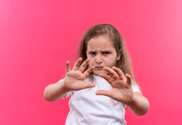 Sad little school girl wearing white t-shirt showing stop gesture on isolated pink background