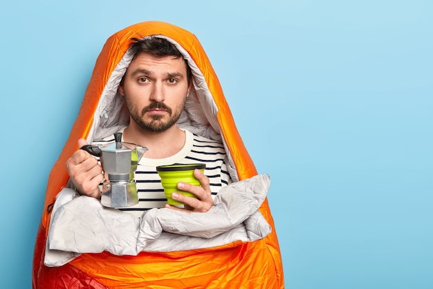 Free Photo sad male camper prepares fresh drink, holds coffee pot, spends free time in nature, enjoys summer morning, wrapped in sleeping bag, stands against blue wall