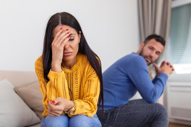 Sad pensive young girl thinking of relationships problems sitting on sofa with offended boyfriend conflicts in marriage