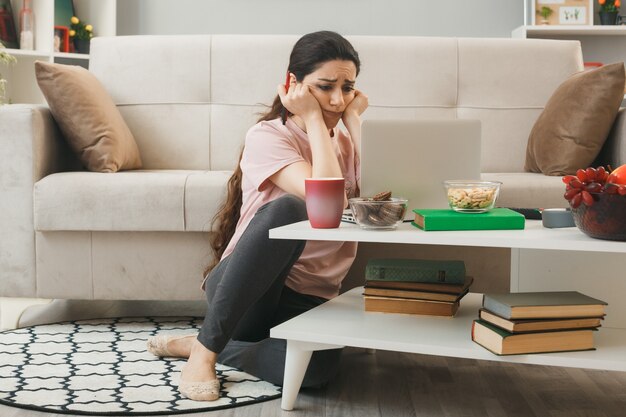 Sad putting hands on cheeks young girl used laptop sitting on floor behind coffee table in living room