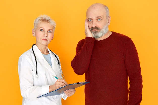 Free photo sad retired female practitioner in white medical coat holding clipboard telling her elderly patient about diagnosis and treatment