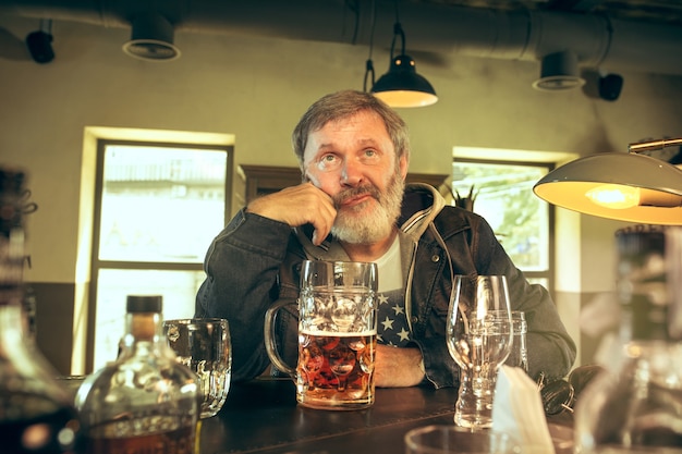 Free photo sad senior man drinking alcohol in pub and watching a sport program on tv