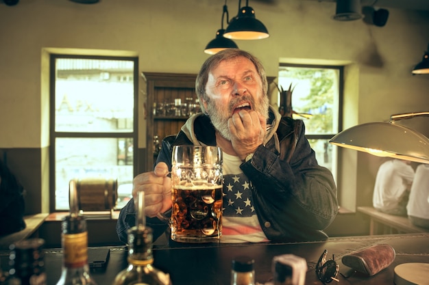 Free Photo sad senior man drinking alcohol in pub and watching a sport program on tv