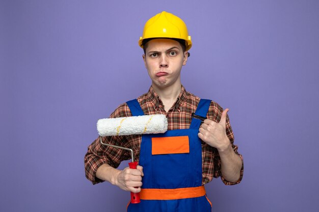 Sad showing thumb up young male builder wearing uniform holding roller brush 