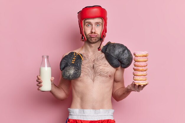 Sad skinny male boxer wears hat and boxing groves around neck holds pile of donuts and bottle of milk.