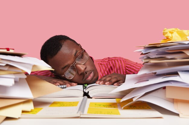 Sad sleepy overworked male scientist leans head on table, being tired of doing scientific work, studies papers, writes in notepad