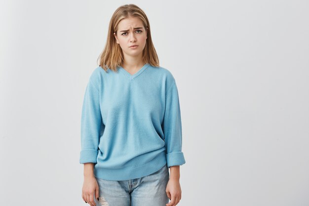 Sad, unhappy, beautiful woman with straight fair hair having dark charming eyes posing at studio  , upset because of bad news. Pretty girl in blue sweater and jeans.