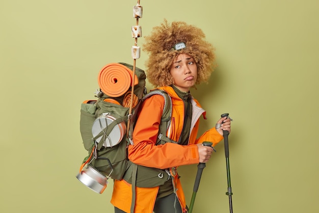 Free photo sad unhappy woman backpacker carries rucksack uses trekking sticks feels tired walks long ditance isolated over green background discontent female hiker going to climb mountain adventure travel