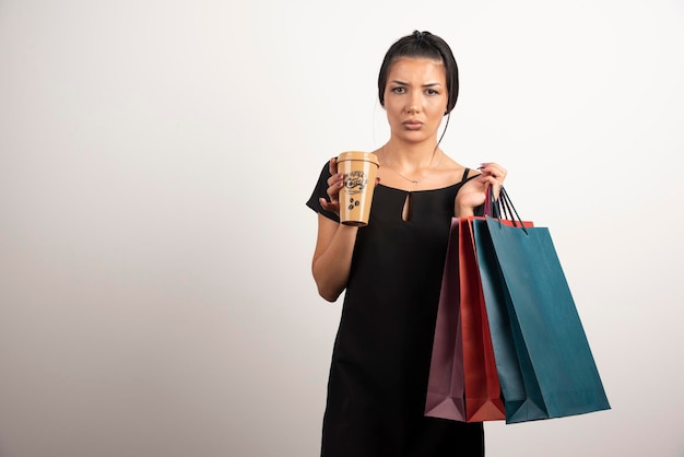 Sad woman carrying shopping bags and coffee on white wall.