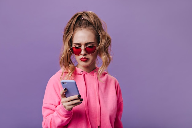 Free photo sad woman in hoodie and red sunglasses reads message in her phone portrait of woman in bad mood holding cell on purple isolated background