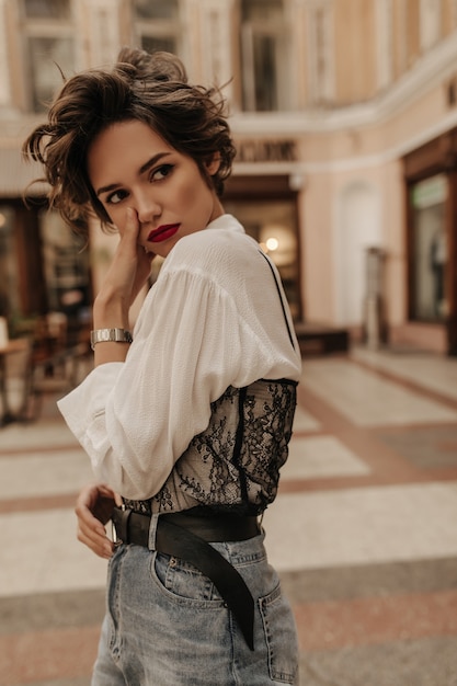 Sad woman in light blouse with lace and jeans with belt posing at street. Trendy woman with short hair and red lipstick looks away in city.